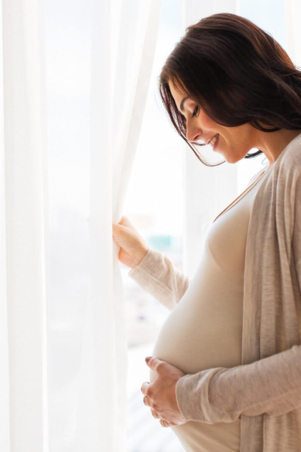 A happy young pregnant woman with a big stomach is standing by the window and caressing her stomach.