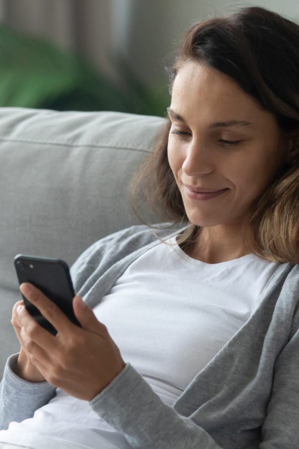 Una donna sorridente si riposa seduta confortevolmente sul divano in soggiorno e naviga in internet con uno smartphone.